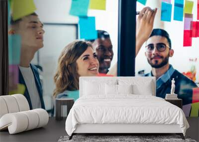 Diverse team of positive young people laughing while working together during brainstorming and standing behind glass wall with sticky colorful papers.Cheerful students learning words from stickers Wall mural