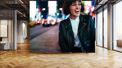 Excited young woman with short haircut laughing at urban setting with night lights enjoying leisure time in New York, happy hipster in stylish wear and eyewear having fun on megapolis street Wall mural