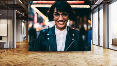 Half-length portrait of cheerful hipster girl in eyewear enjoying night cityscape standing on urban illumination background, gorgeous woman in stylish clothing looking at camera standing on Manhattan Wall mural