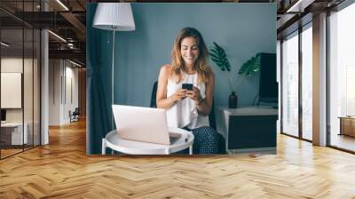 Happy female blogger in casual wear working remotely at home interior enjoying online chatting during break, cheerful Caucasian freelancer networking websites via modern smartphone gadget Wall mural