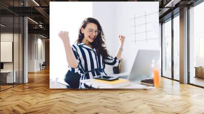 Happy young woman sitting at table with hands up in winner gesture Wall mural