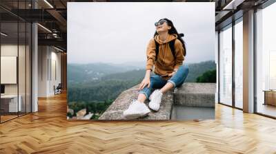 Overjoyed asian female traveler in sunglasses enjoying active leisure and destination exploring, smiling woman tourist sitting outdoors satisfied with wanderlust tour on discovering locations Wall mural