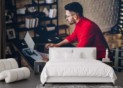 Pensive bearded young man in eyeglasses checking information on website on modern laptop computer using wireless internet.Concentrated graphic designer with sketches in hands working on freelance Wall mural