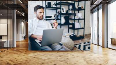 Pensive caucasian man concentrated on information for making shopping from home using laptop computer and wifi, serious hipster guy confused with credit card number making transaction on netbook Wall mural