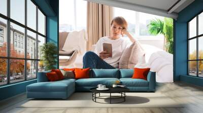 Pensive middle aged woman with smartphone relaxing on sofa Wall mural