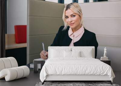 Portrait of beautiful manager holding pen in hand and looking at camera while sitting with books in workshop with modern interior, successful executive female entrepreneur spending time for knowledge Wall mural