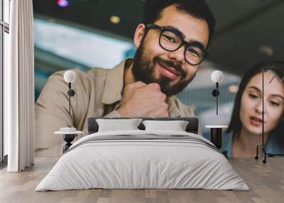 Portrait of cheerful bearded hipster guy in eyewear smiling at camera and enjoying of spending leisure time with beautiful caucasian girlfriend sitting near and looking on screen of cellular phone Wall mural