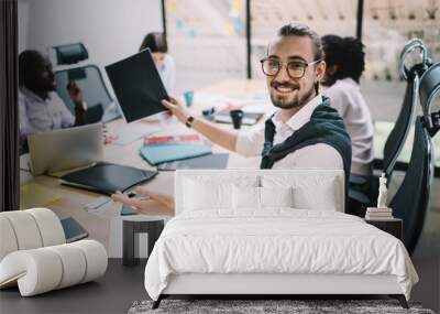 Portrait of cheerful caucasian male employee in optical eyewear for vision correction holding black paper in hand and smiling at camera feeling good from cooperation on information with friendly team Wall mural