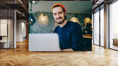 Portrait of cheerful caucasian man in trendy bennie satisfied with online language courses using netbook for studying, happy bearded hipster looking at camera satisfied with wifi for remote work Wall mural