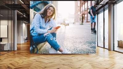 Portrait of cheerful woman in spectacles holding cellphone gadget and smiling at urban setting while resting in city, joyful hipster girl in stylish apparel using mobile phone while posing at street Wall mural