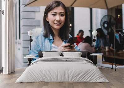 Portrait of happy Chinese female blogger in stylish denim apparel making successfully transferring money via free 4g high speed internet connection on smartphone resting at outdoors cafeteria Wall mural