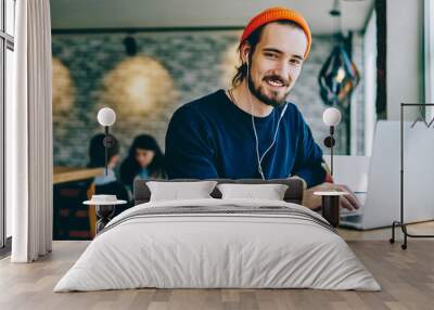 Portrait of happy smiling hipster guy enjoying time for favourite music playlist during e learning in cafeteria, positive male teenager in headphones rest indoors sitting at table with laptop device Wall mural