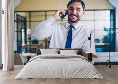 Portrait of positive professional male economist talking with colleague using wireless internet and application on modern mobile phone while sitting at desktop with laptop device in company office Wall mural