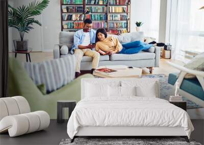 Positive African American couple in love resting together at apartment on comfortable sofa, dark skinned male showing new app on smartphone to his girlfriend lying with book in modern home interior Wall mural