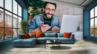 Positive caucasian hipster guy in eyewear laughing at content from social networks using mobile phone for browsing, cheerful male freelancer having fun during remote job  chatting on cellular. Wall mural