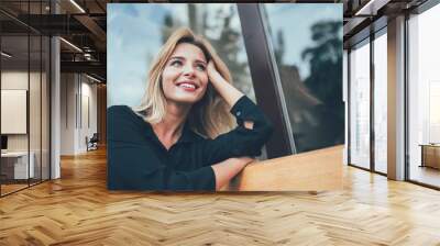 Positive cheerful hipster girl with perfect blonde hair taking rest on bench in city and enjoying time for dreaming outdoors, happy female with cute candid smile looking away during leisure Wall mural