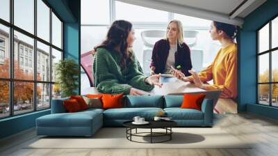 Positive crew of female designers having conversation during cooperation process in coworking office, young woman explaining ideas to her colleague planning strategy sitting next to laptop computer Wall mural