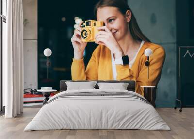 Positive hipster girl casual dressed focusing on vintage camera and making photos indoors, successful amateur photographer taking pictures via old fashioned equipment enjoying hoppy in cafeteria Wall mural