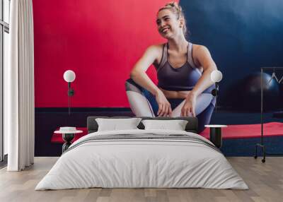 Positive hipster girl dressed in stylish tracksuit smiling while taking rest on red carpet for pilates training, happy female athlete enjoying time for crossfit in sport hall feeling carefree Wall mural