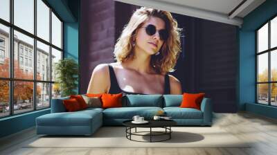 Positive hipster girl in sunglasses waiting friend on urban setting during sunny summer day, attractive caucasian female teenager in stylish outfit standing at publicity area and enjoying leisure Wall mural