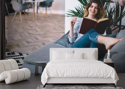 Positive young woman happy with recreation time at home enjoying interesting book with funny plot.Cheerful hipster girl lying on comfortable couch and reading textbook in modern apartment Wall mural