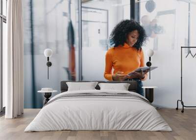 Professional Black businesswoman using tablet in modern office setting, focused and serious, wearing casual orange sweater and jeans, exemplifying mobile business management. Wall mural