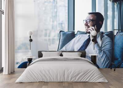 Side view of happy man in eyewear looking away and smiling while communicating with friend via smartphone gadget sitting in office space with laptop computer, concept of business leaders - people Wall mural