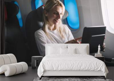 Smiling attractive female passenger searching movie to watch on board tv device during comfortable flight. Caucasian young woman traveler enjoying touch screen entertainment monitor system on airplane Wall mural