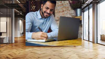 Smiling attractive male economist making financial report counting amount of sales using information from corporative database working hard in coworking space using modern laptop computer and wifi Wall mural