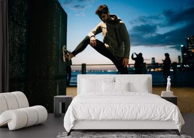 Sportsman dressed in tracksuit doing stretching while enjoying music during evening workout in urban setting.Young man in sport wear doing exercise training outdoors while listening audio in earphones Wall mural