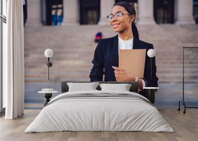 Successful African American graduate juridical specialist standing outdoors against courthouse building with folder in hands. Prosperous female student of high economic university in elegant wear Wall mural