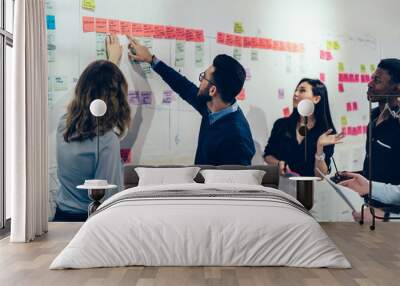 Team of multicultural young people pointing on wall with glued colorful paper notes with foreign words during productive lesson.Diverse group of male and female employees in formal wear using stickers Wall mural