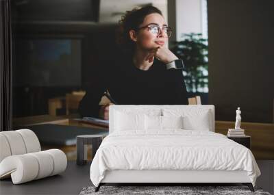 Thoughtful young author businesswoman and thinking on writing interesting article.Woman in stylish eyeglasses with good lenses looking out of window and pondering new plans while noting them Wall mural