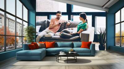 Young marriage spending time together in modern apartment with stylish interior.Young man searching website on smartphone device using home internet while sitting on sofa with girlfriend in flat Wall mural