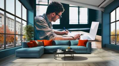 Young millennial male student searching information for homework using laptop and wifi connection indoors, man writing article in textbook for add interesting content on own website or web page Wall mural