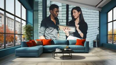 Young woman talking with african american colleague and showing video presentation on website on modern tablet using 4G internet.Two multicultural employees discussing financial news on website Wall mural