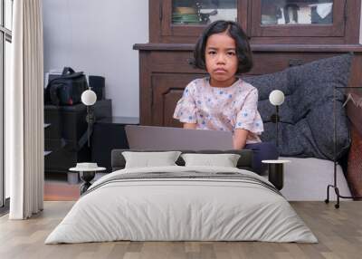 A little girl sitting on the sofa doing her homework on the laptop; upset, unhappy, frowning. Wall mural