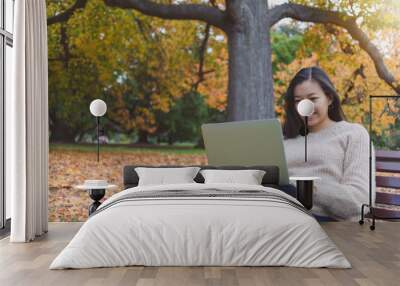 Asian beautiful smiling woman working with laptop while sitting in garden with green grass and falling leaf in autumn.Concept of people using technology. Wall mural