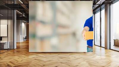 Asian happy delivery man wearing a blue shirt carrying paper parcel boxes isolated on blur interior warehouse in the shopping mall background.Concept of Postal delivery service. Wall mural