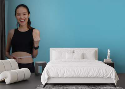 Portrait of confident beautiful asian fitness woman standing and use tape measure to measure waist  after exercise with smiling face on blue colour background.Concept of slim and healthy happy girl. Wall mural