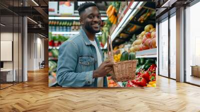 A close-up of a happy, stylish young man shopping in the fresh produce section of a supermarket, holding a basket while browsing colorful fruits and vegetables displayed in the grocery store. Wall mural