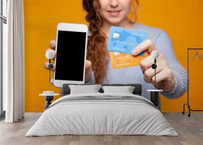 Girl holding white iphone and two gredit cards isolated over the yellow background Wall mural