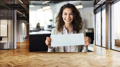 Smiling businesswoman holding a cheque, standing in a modern office setting, symbolizing financial success and business achievements. Wall mural