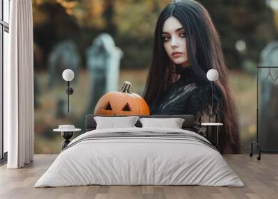 Beautiful goth girl with a long lush hair wearing black gothic dress, holding a halloween decoration pumpkin. Graveyard cemetery blurred background. Ready for Halloween gothic party Wall mural