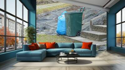 one green plastic trash can and a blue plastic bag with garbage stand on green grass near gray concrete steps outside Wall mural