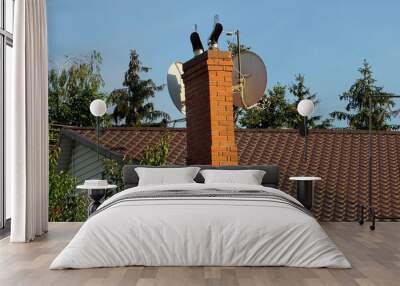 one red brick chimney with two white satellite dishes on a brown tiled roof against a blue sky Wall mural