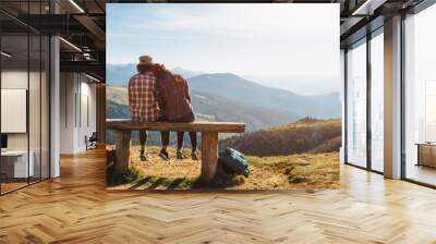 Couple of hikers with backpacks enjoying valley landscape view from top of a mountain. Young adult tourists, man and woman sitting in the bench. Panoramic view of mountain hills, Carpathian mountains Wall mural