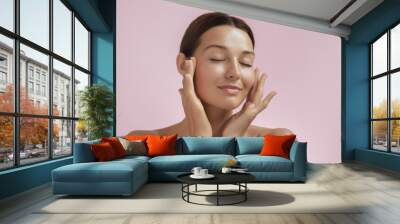 Skincare and Beauty Portrait. Woman with beautiful face touching healthy facial skin. Young adult smiling Asian girl model with natural makeup touching glowing hydrated skin on pink background closeup Wall mural