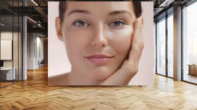 Skincare. Woman with beautiful face touching healthy facial skin. Fascinating portrait of confident girl model with natural makeup enjoys glowing hydrated skin on beige background closeup. High Wall mural