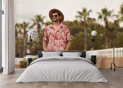 Tourist man on the beach standing outdoors against resort with palm trees in the background during summer holidays. The guy wearing a summer casual shirt hat and sunglasses, standing looking to the Wall mural